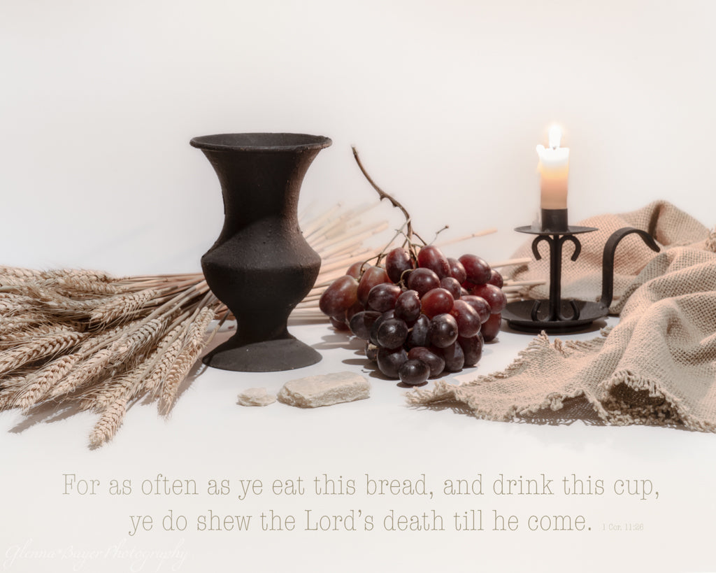 Black drinking cup, grapes, communion bread, and candle on a white surface with a Bible verse.