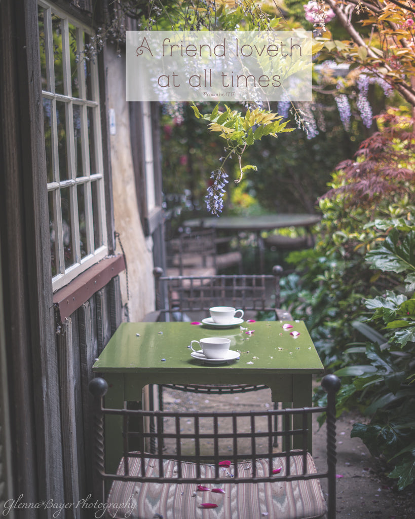 Green table with tea cups in a garden setting next to a windowed wall, surrounded by plants and flowers.