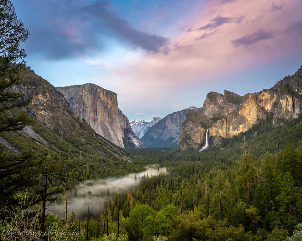 Valley view with mountains, trees, and a waterfall under a pink and blue sky.