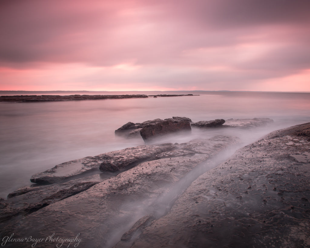Pink sunrise and ocean flowing over rocks in Jervis Bay, Australia